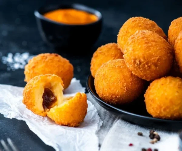 A close-up of crispy fried cheesey chutney bite on a black plate, with one broken open to reveal a gooey melted cheese and dark filling inside. A small bowl of dipping sauce is blurred in the background.