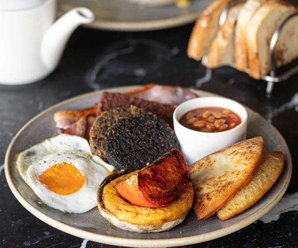 A hearty breakfast plate featuring a fried egg, grilled tomato, potato rosti ring, black pudding, bacon, and a small bowl of baked beans, served on a dark marble table with toast and tea in the background.